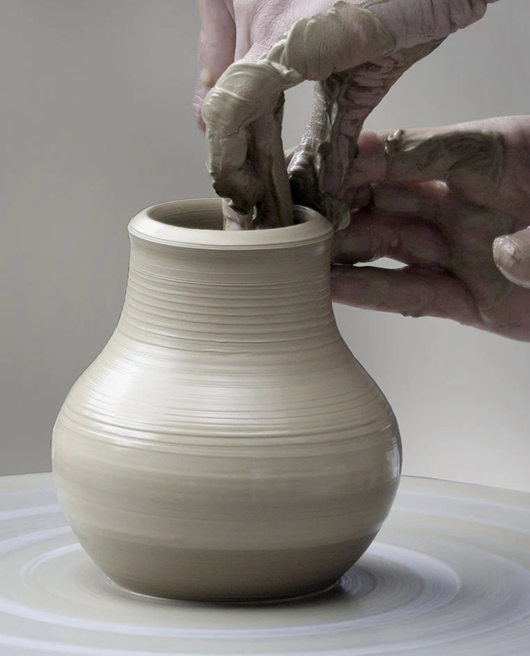A grey clay vase being shaped on throwing wheel in front of grey background